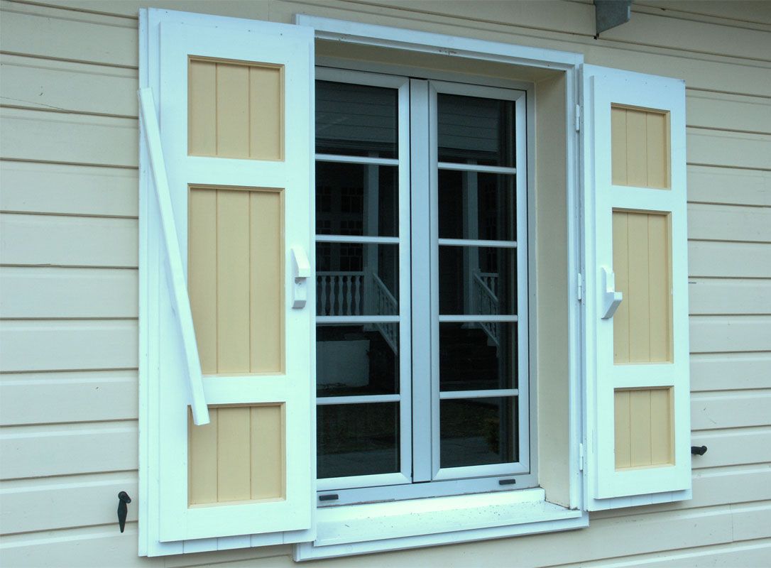 Une fenêtre à cadre blanc et six carreaux, flanquée de deux volets en bois couleur crème, dont l'un est légèrement de travers.