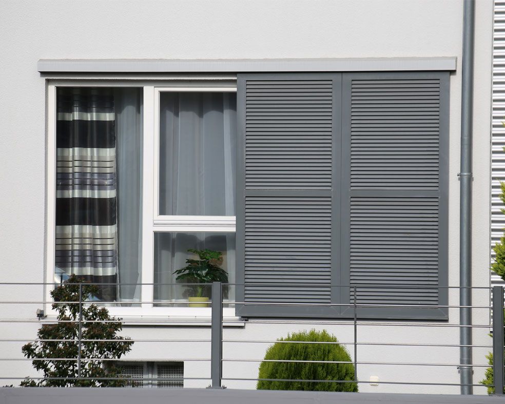 Une fenêtre à cadre blanc, à moitié recouverte de volets coulissants gris foncé, donnant sur une balustrade métallique et de la verdure.