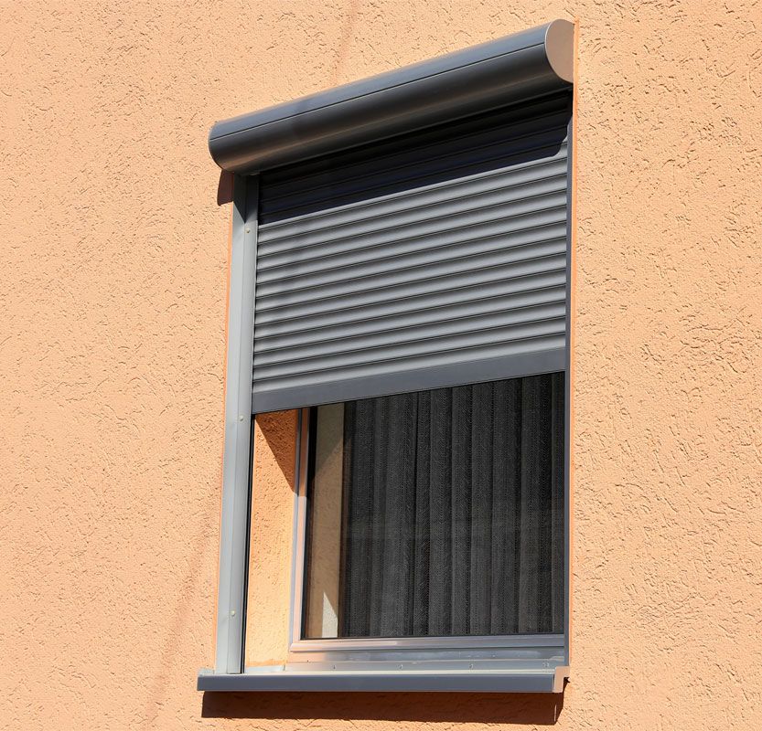 Un volet roulant extérieur gris partiellement abaissé sur une fenêtre donnant sur un mur en stuc couleur pêche.