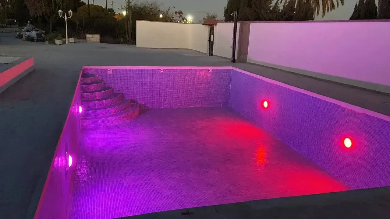 Piscina rectangular vacía al anochecer, iluminada por luces subacuáticas de color púrpura y rojo vibrantes.