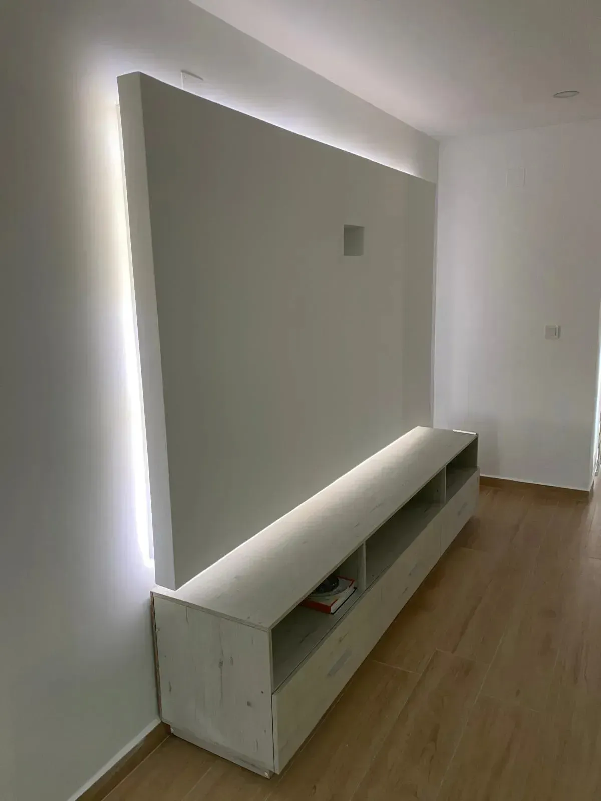 Mueble de TV mural con retroiluminación LED, paneles blancos y consola de almacenamiento abierta sobre suelo de madera clara.