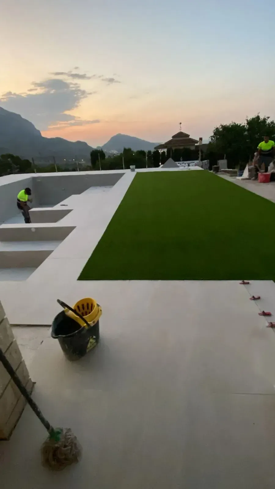 Al atardecer, unos trabajadores terminan de acondicionar un patio y una zona de césped junto a una piscina vacía.