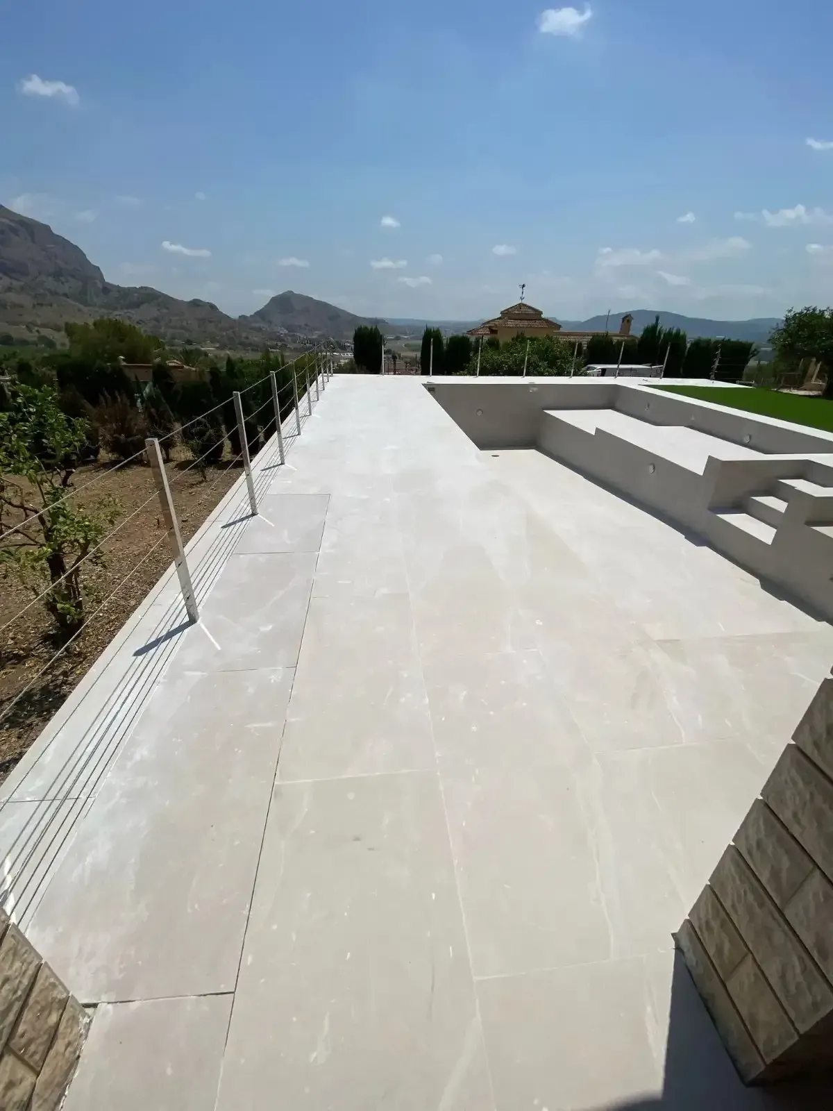 Un patio de piedra de color claro conduce a un banco escalonado y a una zona de césped, con vistas a un paisaje.