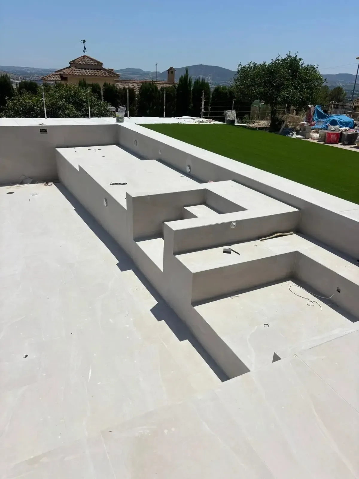 Una piscina vacía de hormigón de color claro con escalones escalonados, vista desde arriba, junto a un césped.