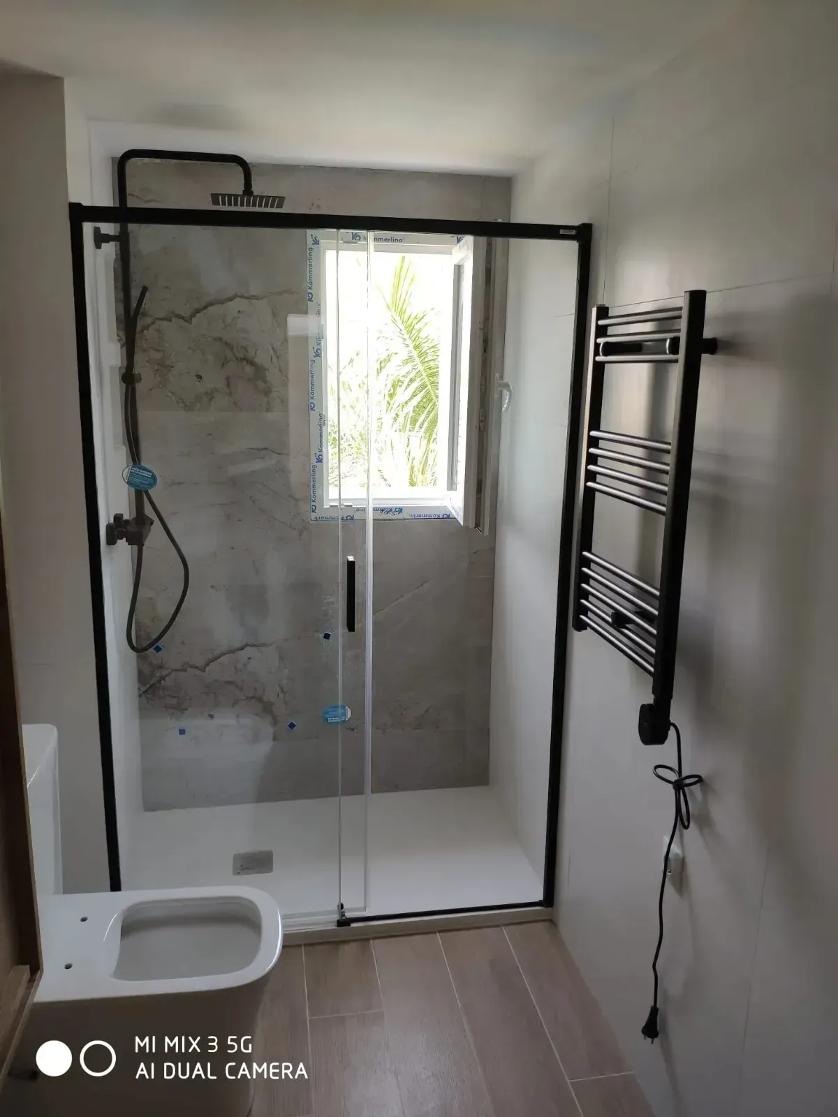 Un baño moderno con ducha acristalada, toallero eléctrico de pared y un inodoro blanco sobre un suelo de baldosas de madera.