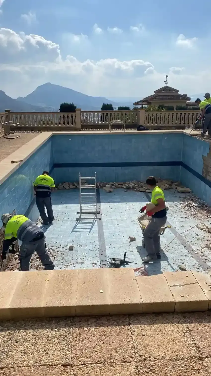 Tres trabajadores con chalecos reflectantes retiran los azulejos de una piscina vacía con revestimiento.