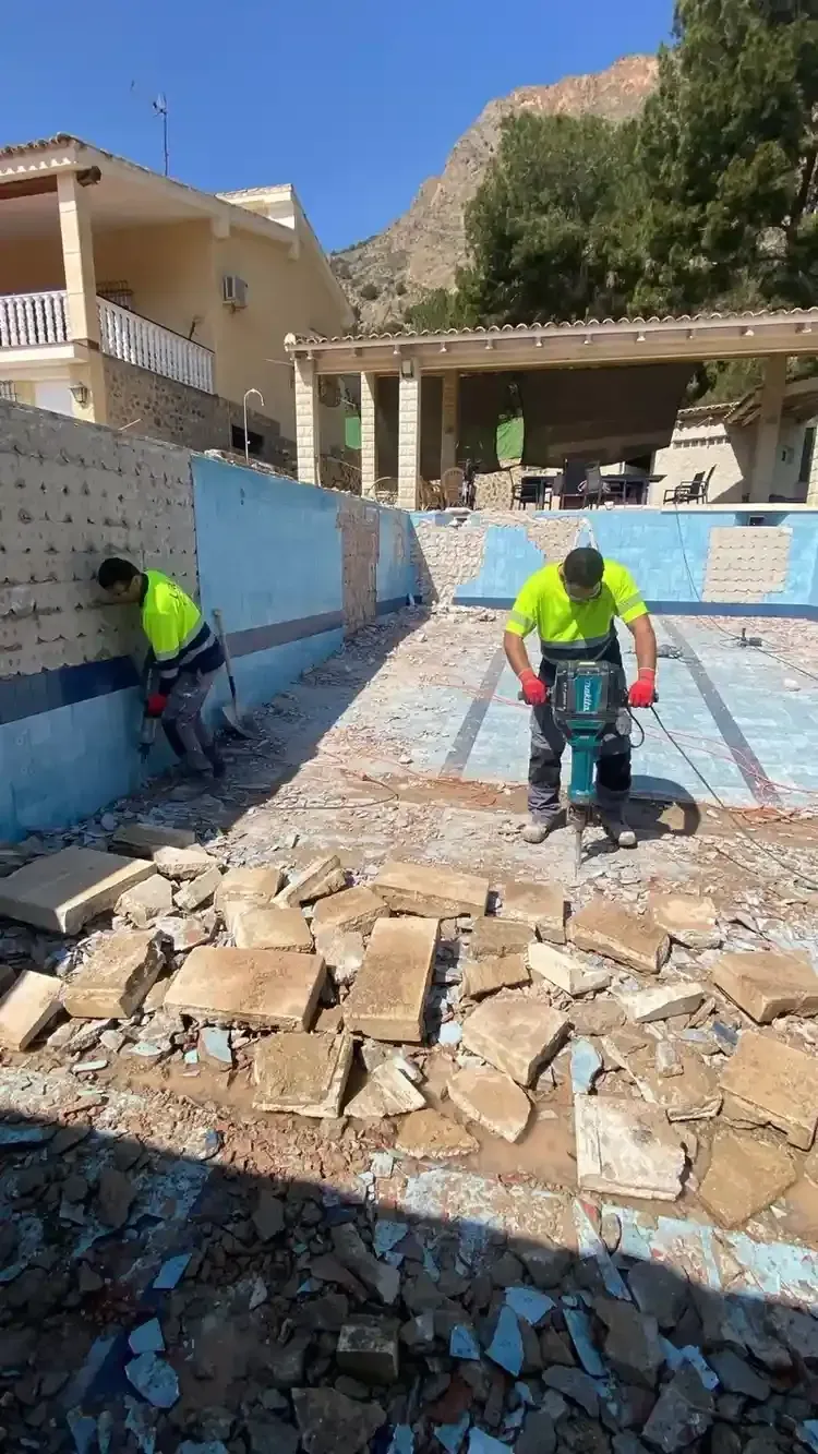 Dos trabajadores con chalecos reflectantes demuelen una antigua piscina de hormigón utilizando martillos.