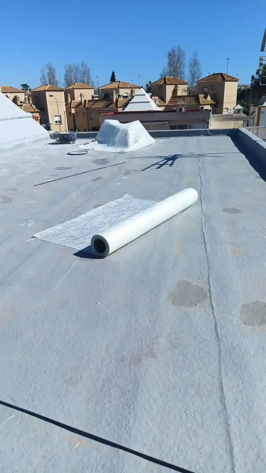 Un rollo de membrana impermeabilizante blanca reposa sobre una azotea industrial plana y gris bajo un cielo azul despejado.