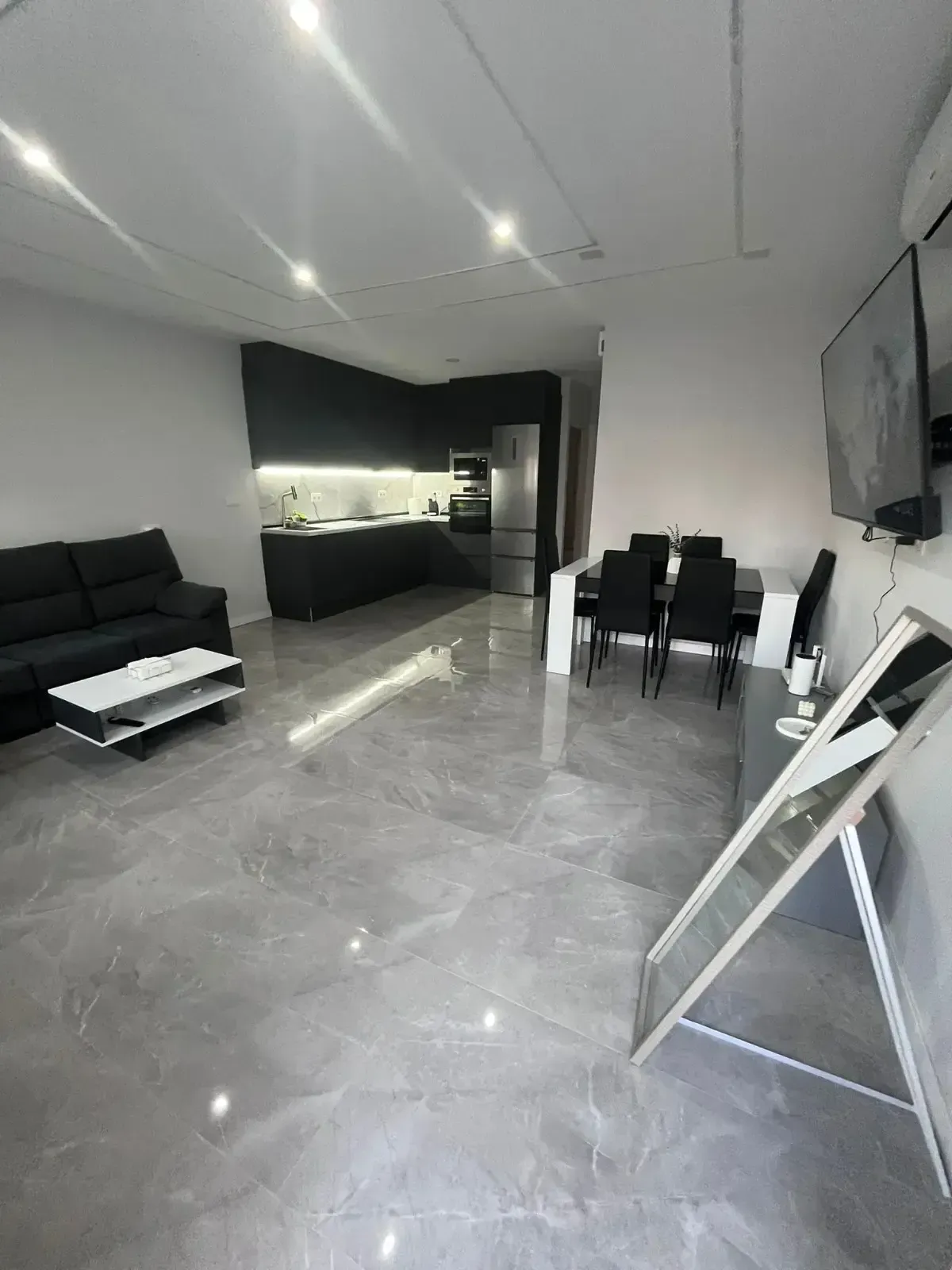 Un moderno salón diáfano con suelo de mármol gris, un sofá negro, una pequeña mesa de centro y una zona de cocina negra.