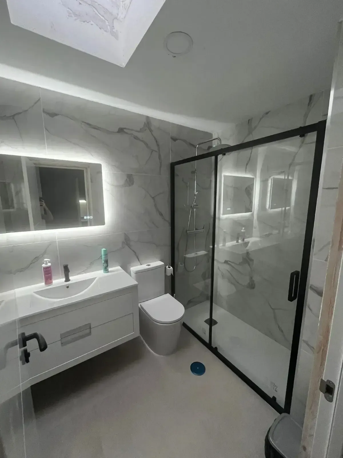 Un baño moderno con paredes con diseño de mármol, un tocador blanco suspendido, un espejo iluminado.