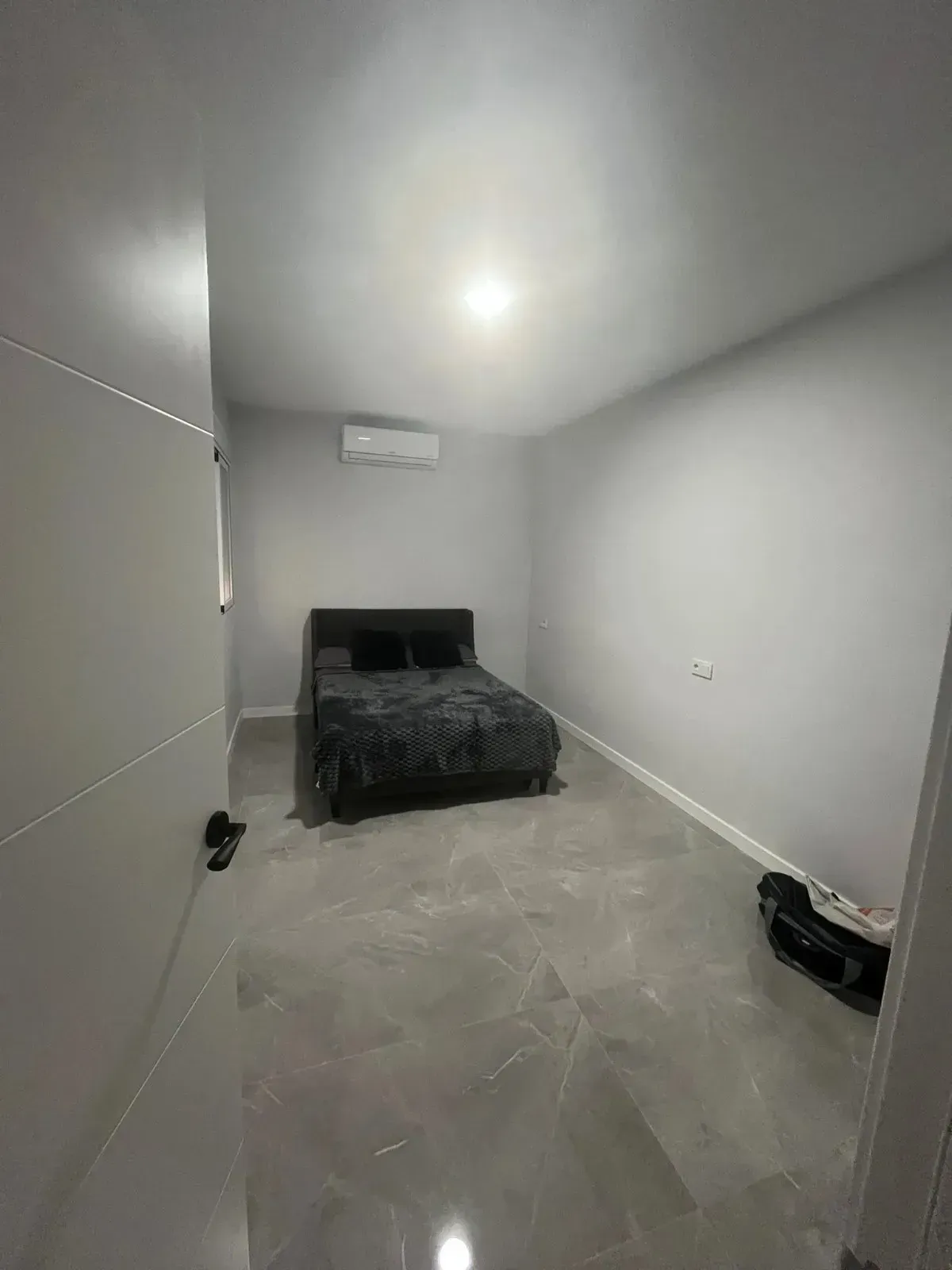 Un dormitorio con mobiliario escaso, con una cama oscura, suelo de baldosas grises, paredes blancas.