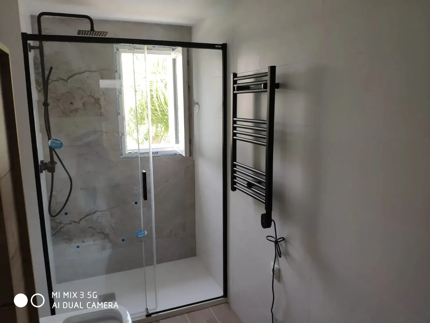 Un baño moderno con ducha con mampara de cristal, grifería negra, interior alicatado, una pequeña ventana.