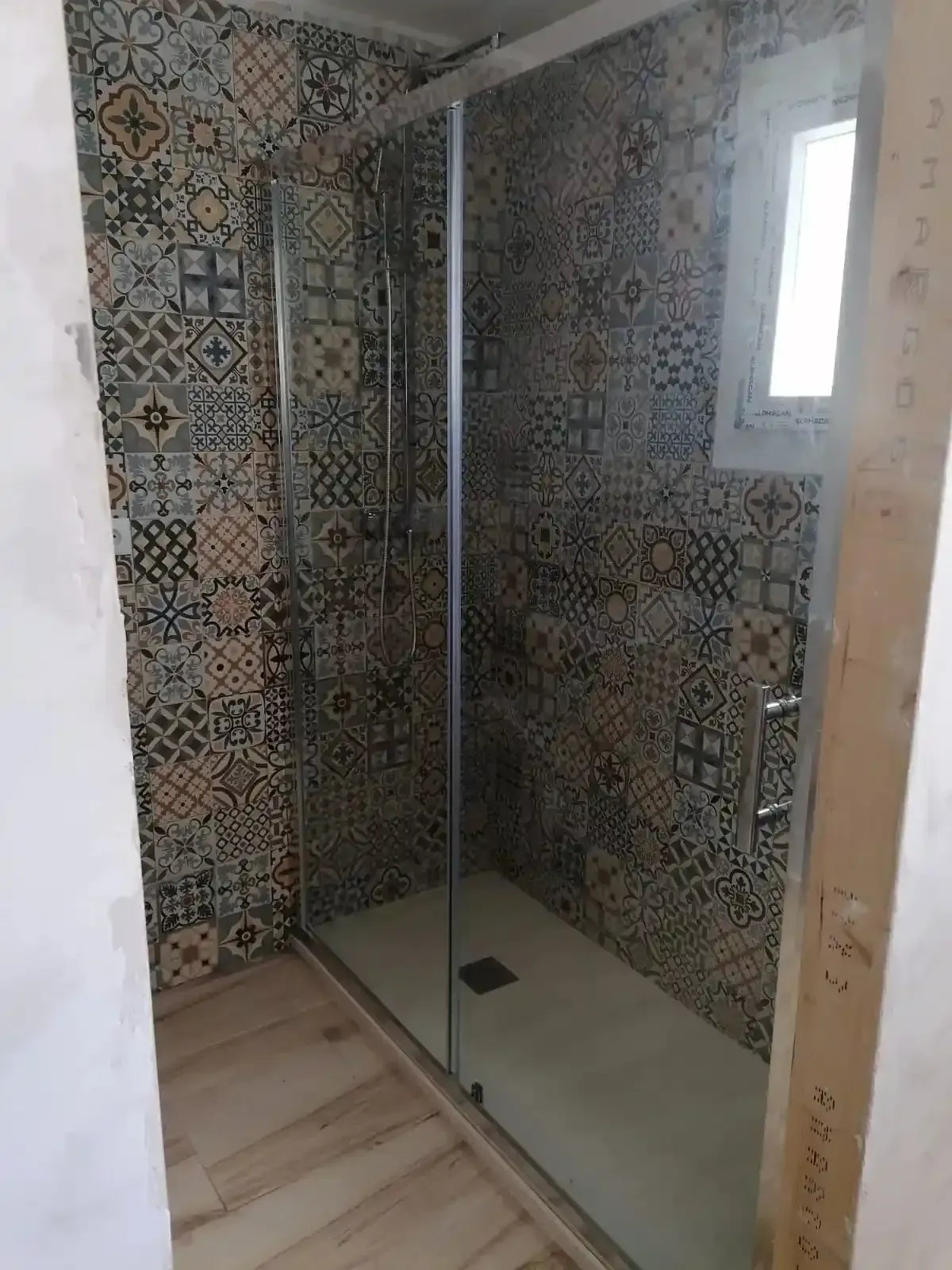Una moderna ducha a ras de suelo con azulejos estampados y multicolores, puerta de cristal y suelo de imitación madera clara.