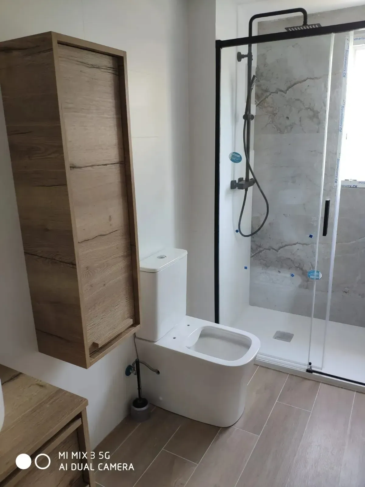 Baño moderno con inodoro blanco, mueble de pared de madera y ducha a ras de suelo con grifería negra.