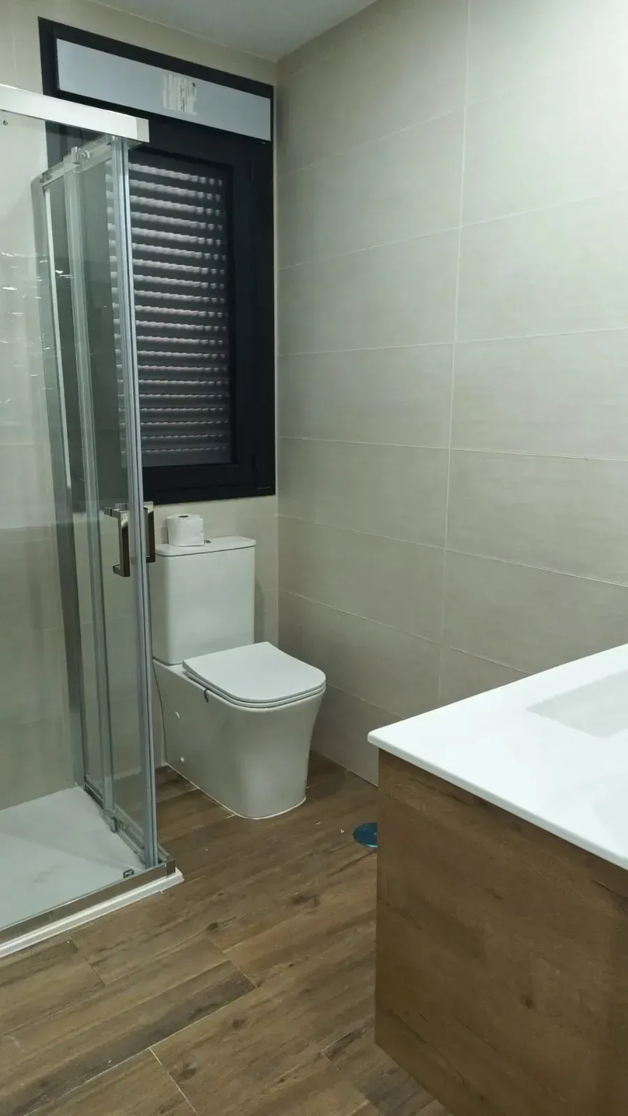 Un baño moderno con suelo de imitación madera, cabina de ducha de cristal, inodoro blanco y tocador con encimera blanca.