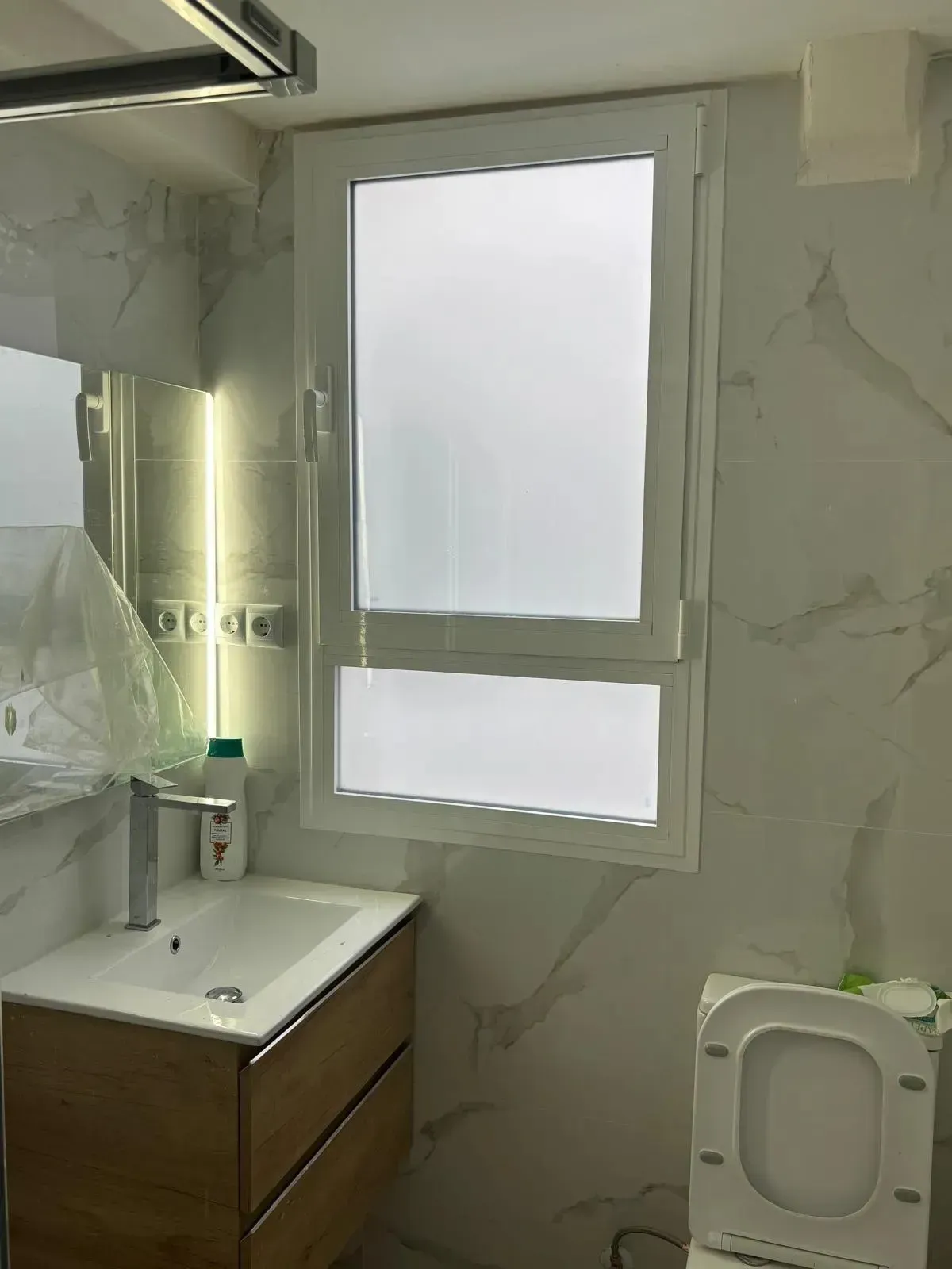 Un baño moderno con paredes de mármol, un mueble de tocador de madera, un lavabo blanco y una ventana de vidrio esmerilado.