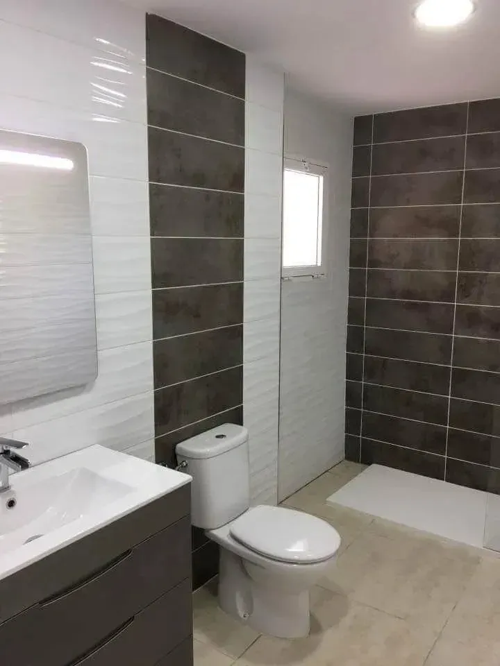 Un baño moderno con lavabo blanco, inodoro y ducha a ras de suelo, con paredes revestidas de azulejos.