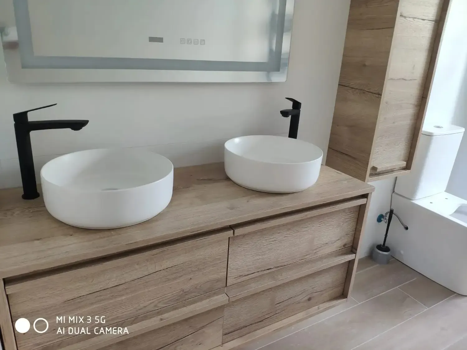 Un moderno mueble de baño con dos lavabos blancos sobre encimera y grifería negra sobre un mueble de madera clara.