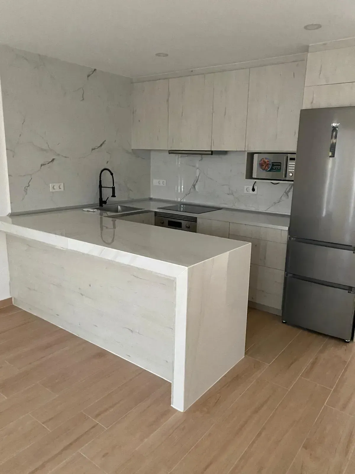 Una cocina moderna con armarios de madera clara, encimeras de mármol blanco, una isla central .