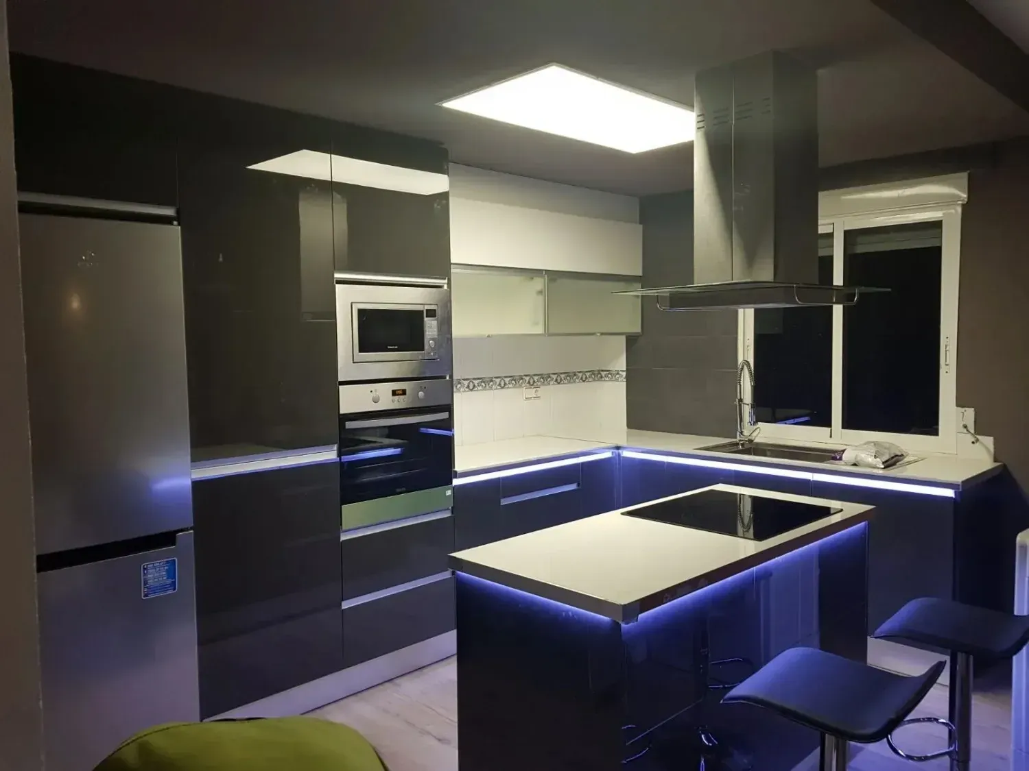 Una cocina moderna con armarios gris oscuro, encimeras blancas, una isla central con iluminación LED azul y dos taburetes.