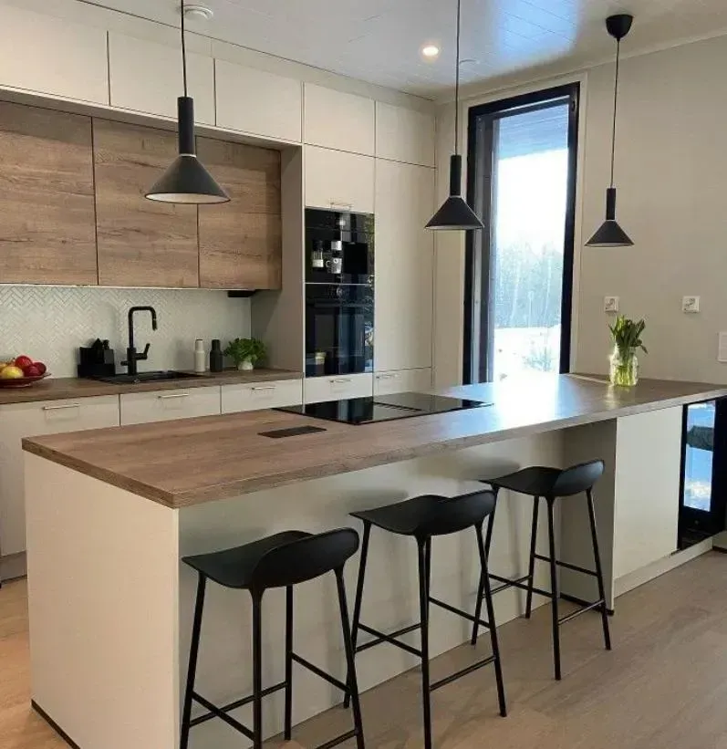 Cocina moderna con armarios claros, una isla con acabado en madera y tres taburetes negros, e iluminación colgante.