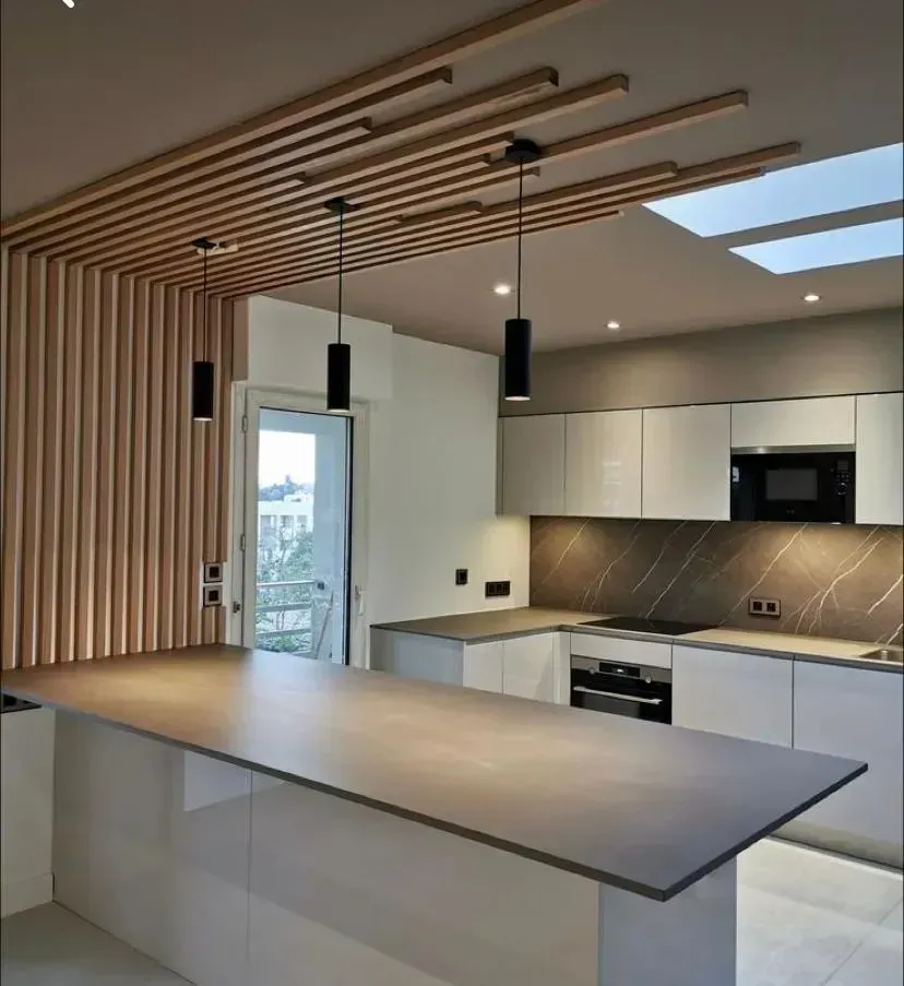 Cocina moderna con techo de listones de madera, armarios blancos, encimera oscura y tres lámparas colgantes negras.