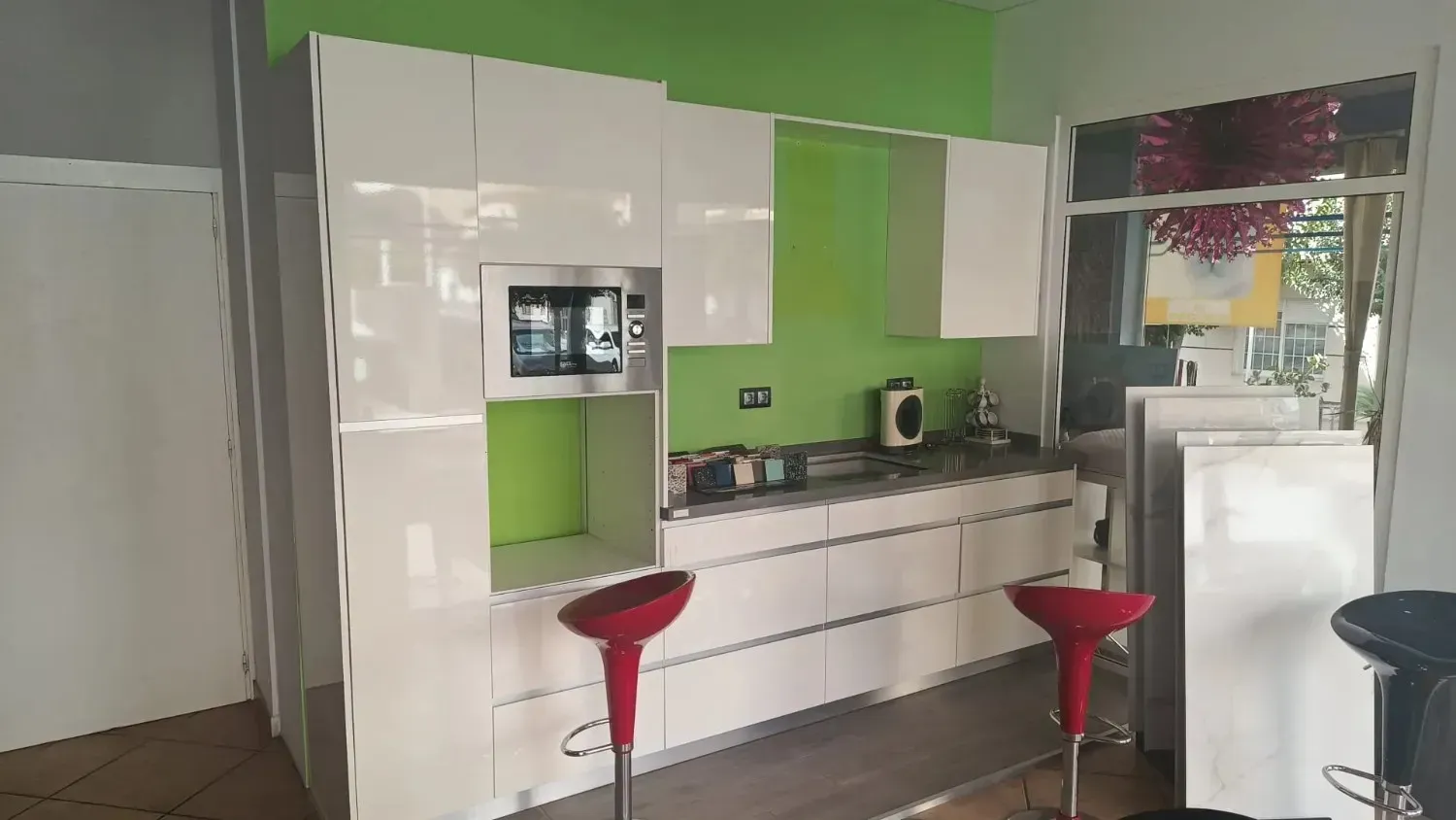 Una cocina moderna con armarios blancos, un salpicadero de color verde brillante, microondas empotrado y dos taburetes rojos.