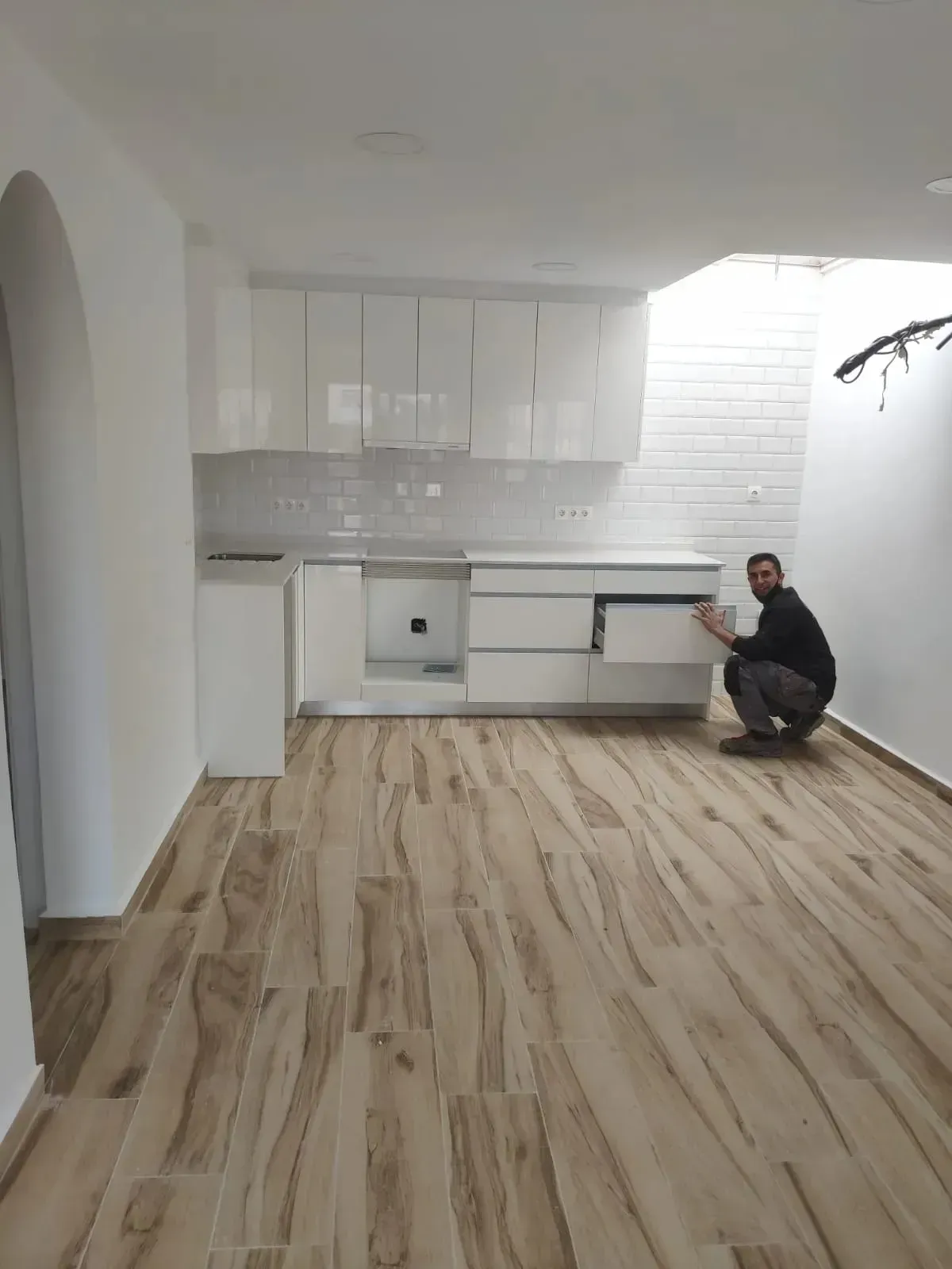 Una persona se agacha para abrir un cajón en una moderna cocina blanca con suelo de baldosas que imitan la madera.