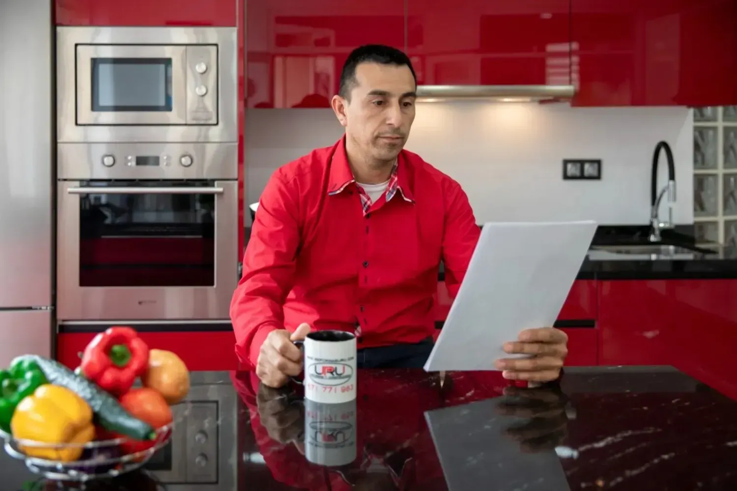 Una persona con una camisa roja está sentada en una cocina roja, sosteniendo una taza y leyendo una tableta.