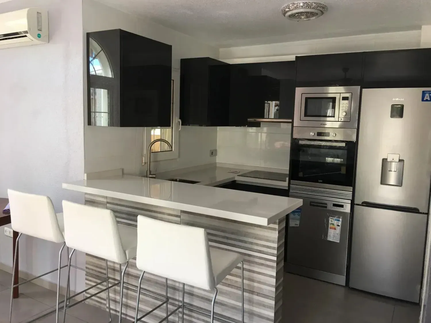 Cocina moderna con barra de desayuno blanca, tres taburetes blancos, armarios negros y electrodomésticos de acero inoxidable.