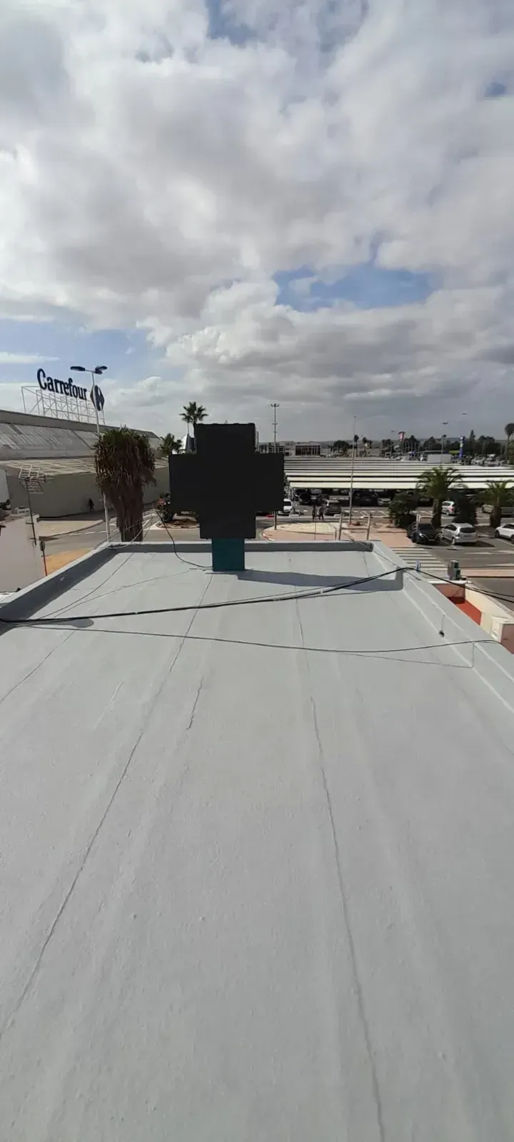 Un tejado plano de color gris con una gran cruz oscura en el centro, contra un cielo azul nublado y un paisaje urbano lejano.
