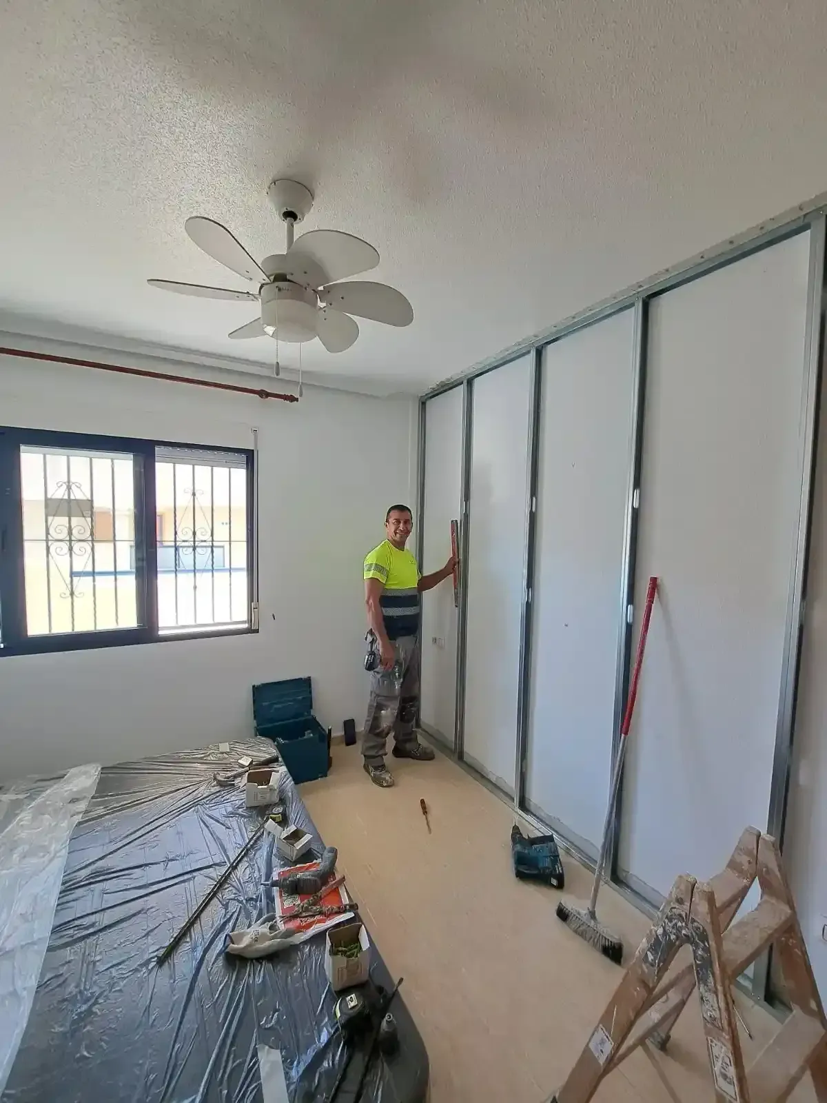 Un trabajador con una camisa amarilla instala la estructura metálica de una pared en una habitación.