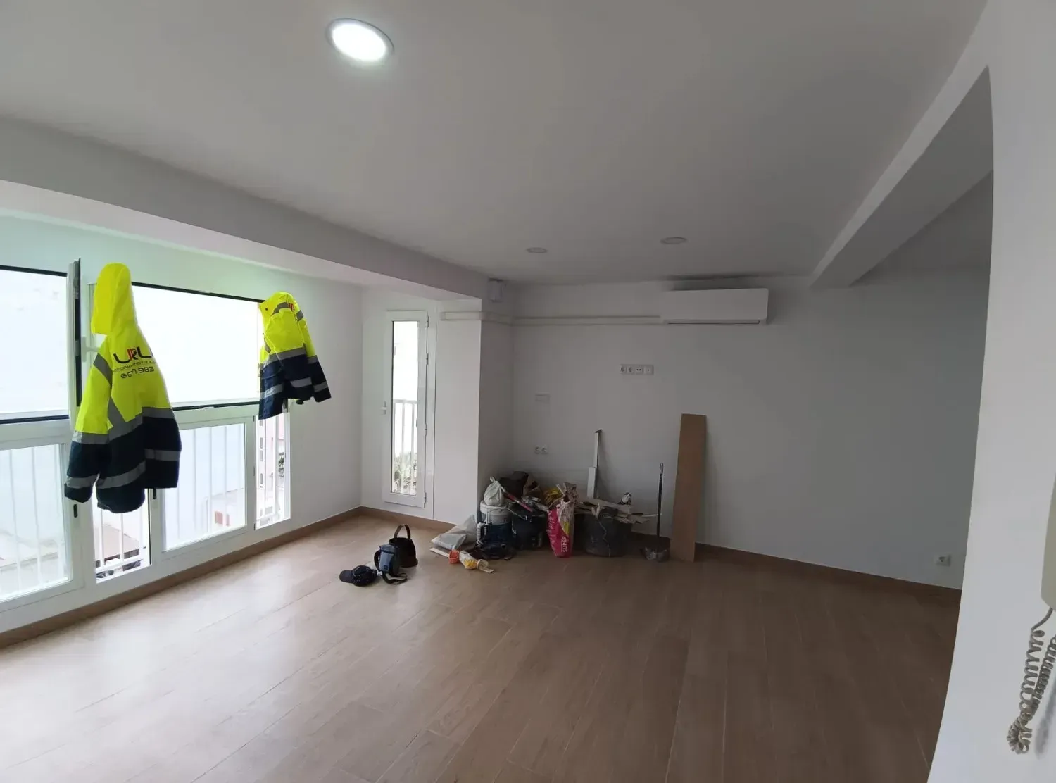 Una habitación vacía, de color claro, con suelo de madera, una ventana con chalecos de seguridad colgado.