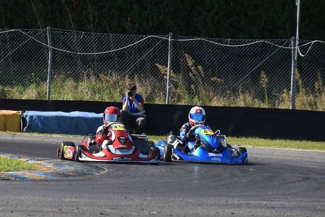 Zwei Go-Karts liefern sich ein Rennen auf der Bahn; rote und blaue Karts treten gegeneinander an. Eine Person beobachtet das Geschehen in der Nähe des Zauns.