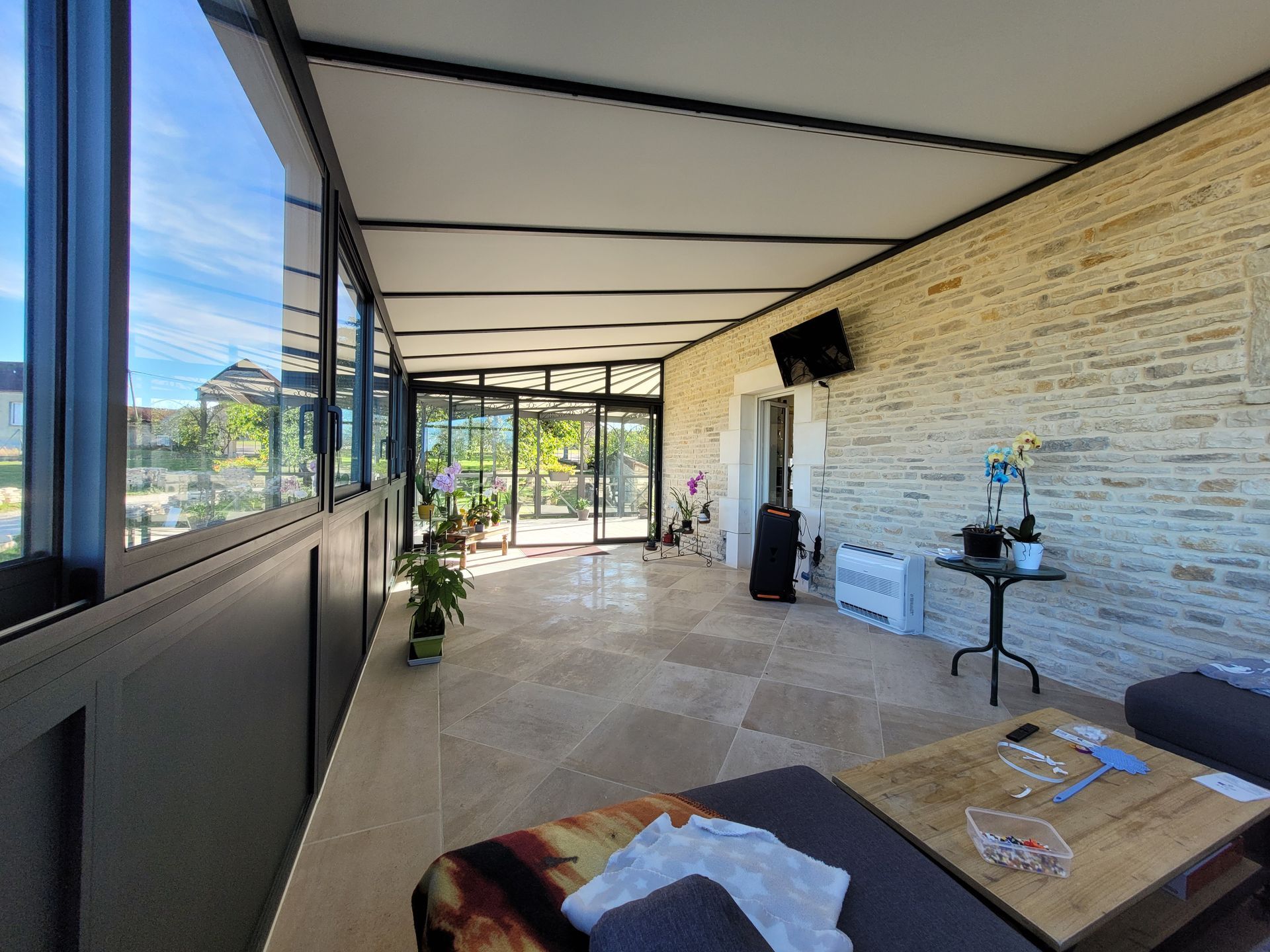 Véranda avec mur en pierre, fenêtres en verre et sol carrelé beige