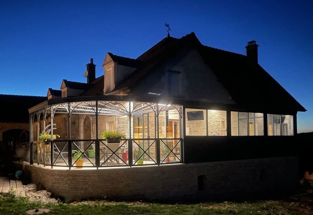 Vue nocturne d'une maison en pierre avec une véranda éclairée aux parois de verre et une balustrade extérieure. Le ciel est d'un bleu foncé