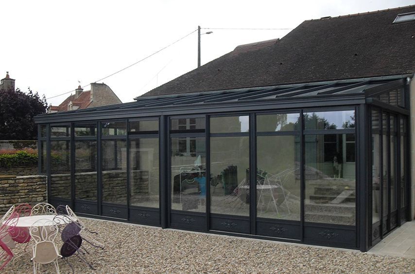 Extension de véranda avec murs et toit en verre, attenante à une maison avec terrasse en gravier et mobilier de jardin