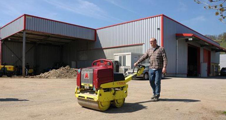 Un homme marche dans la cour d'un entrepôt à côté d'une machine de chantier compacte rouge et jaune.