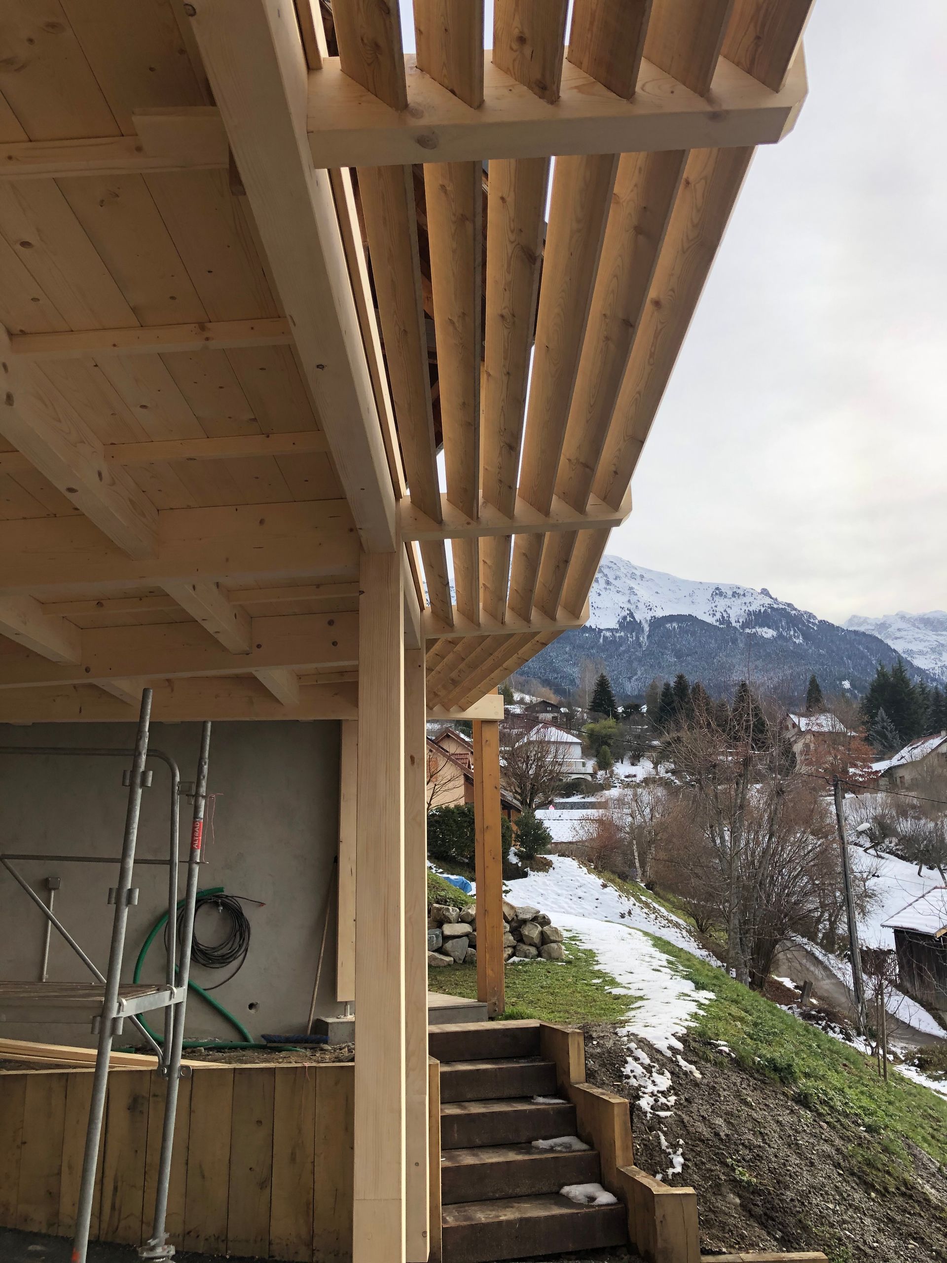 Installation en cours d'un abri de voiture avec une avancée du toit, composé de planches en diagonal avec une légère ouverture entre.