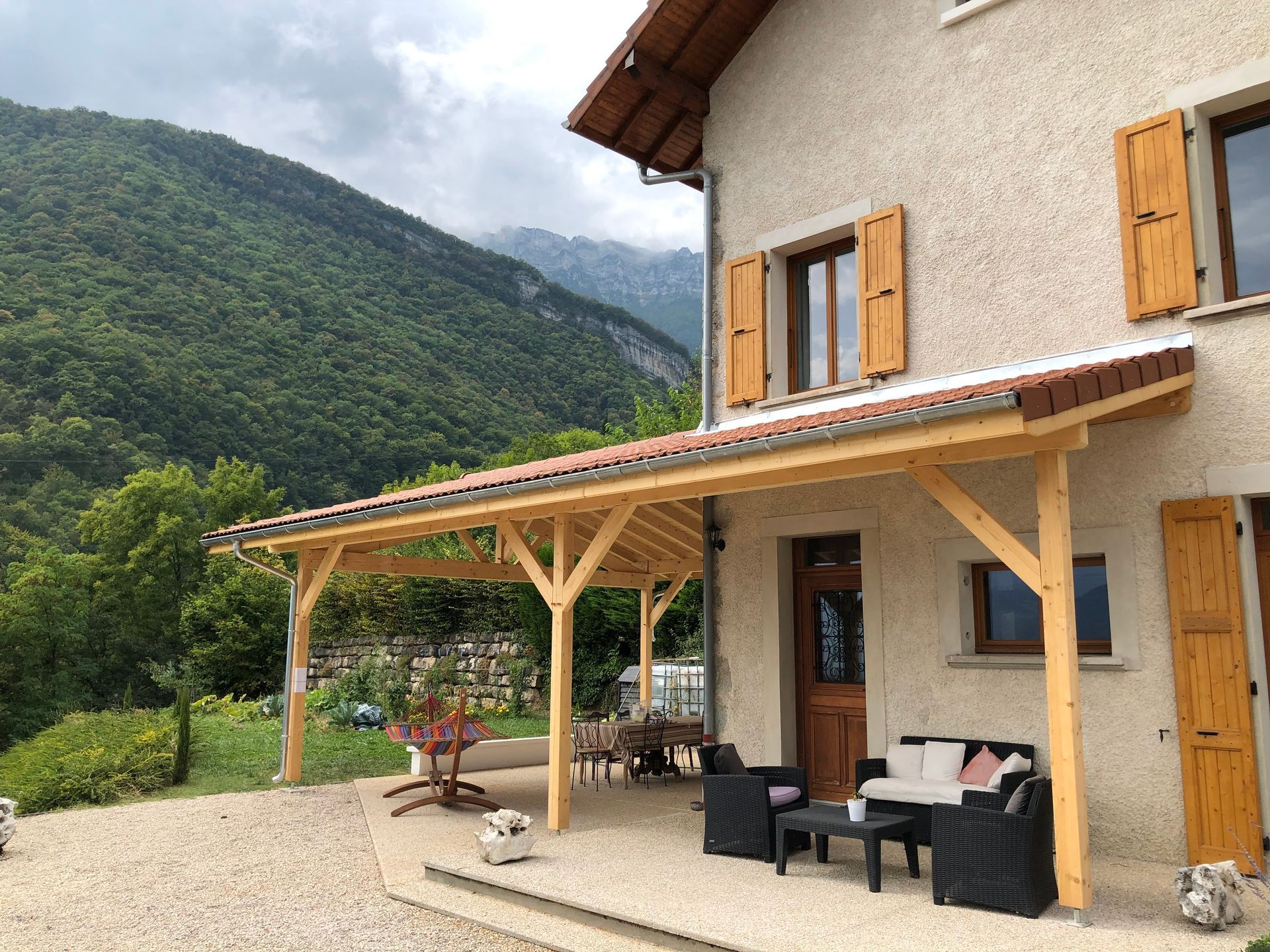 Vue d'un autre angle d'un préau construit au-dessus d'une terrasse,  le long d'une façade, au-dessus d'un salon de jardin.