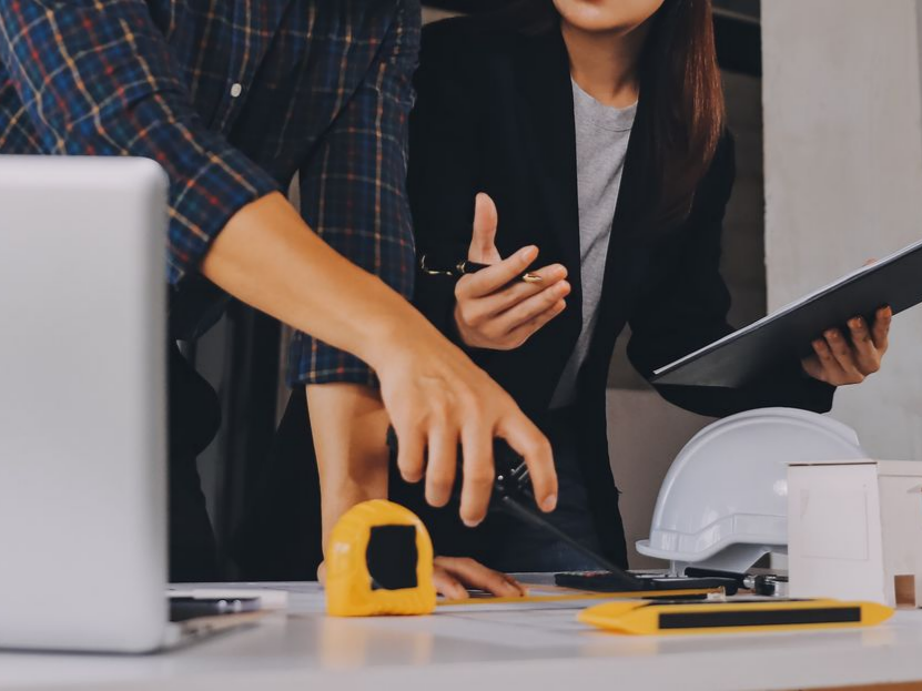 Deux personnes munies d'un mètre ruban, d'un ordinateur portable et d'un casque de sécurité sur un bureau.