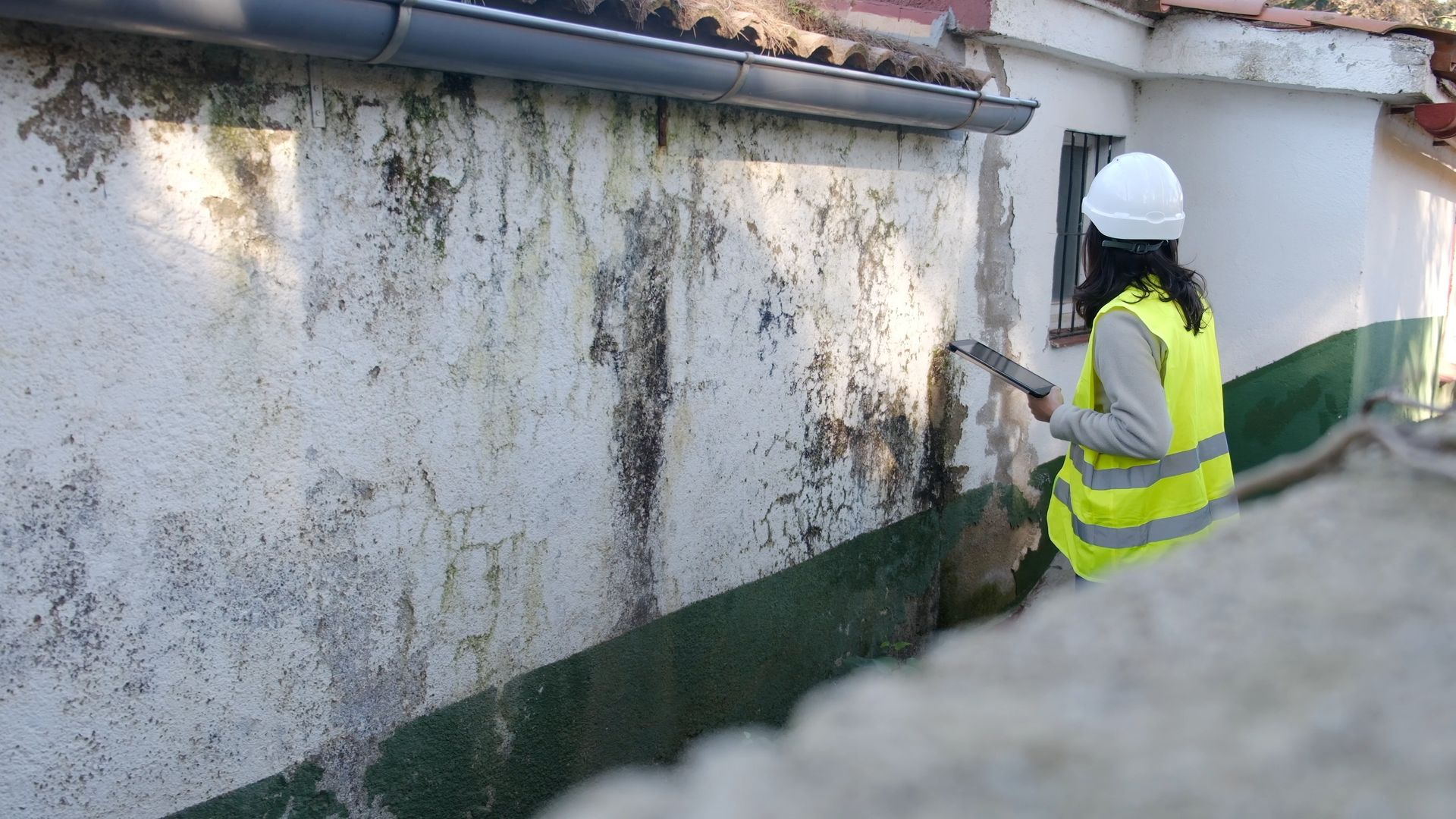 Une femme portant un gilet jaune et un casque de chantier inspecte un mur extérieur blanc taché.