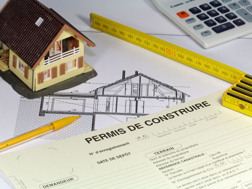 Une maison miniature, un plan, un mètre ruban jaune, un crayon et une calculatrice posés sur une table.