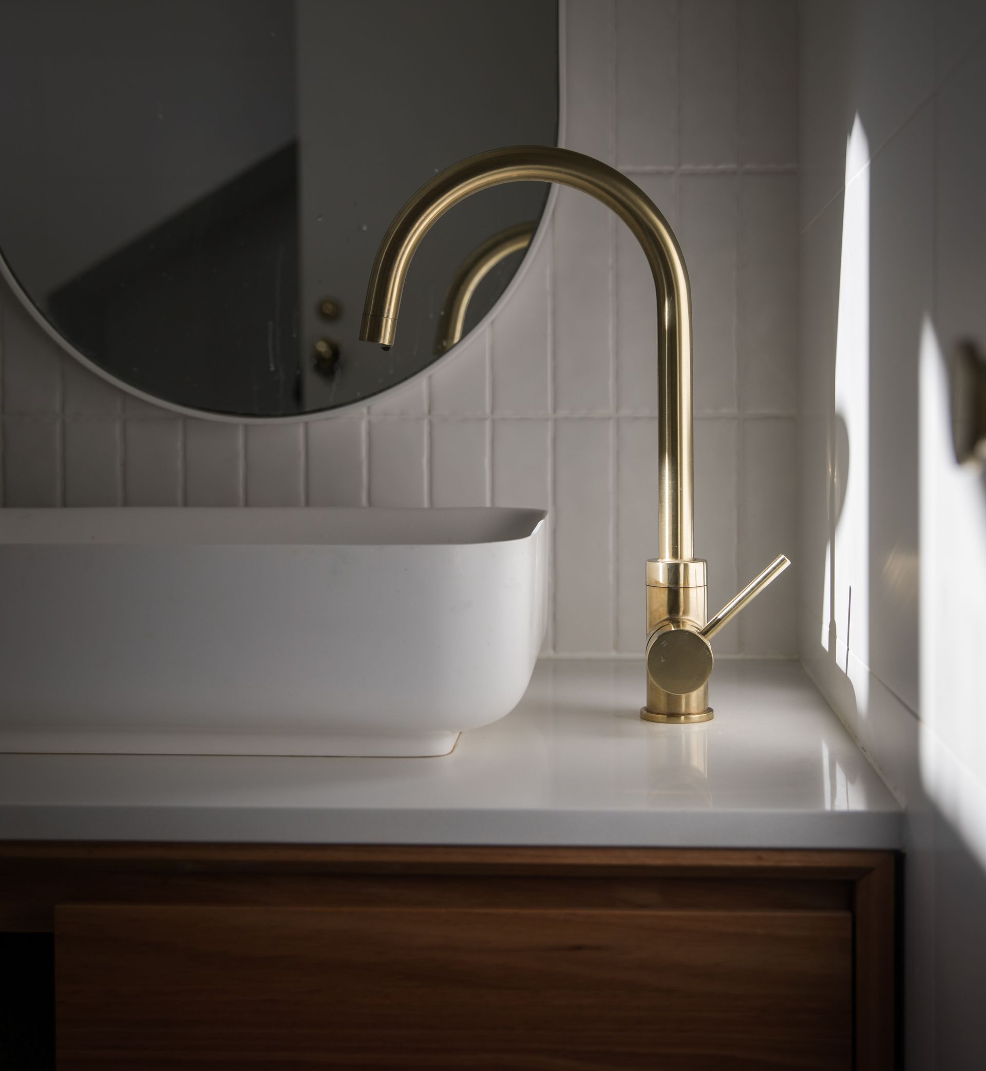 Robinet doré au-dessus d'un lavabo rectangulaire blanc, sur un meuble en bois, avec un miroir rond en arrière-plan.