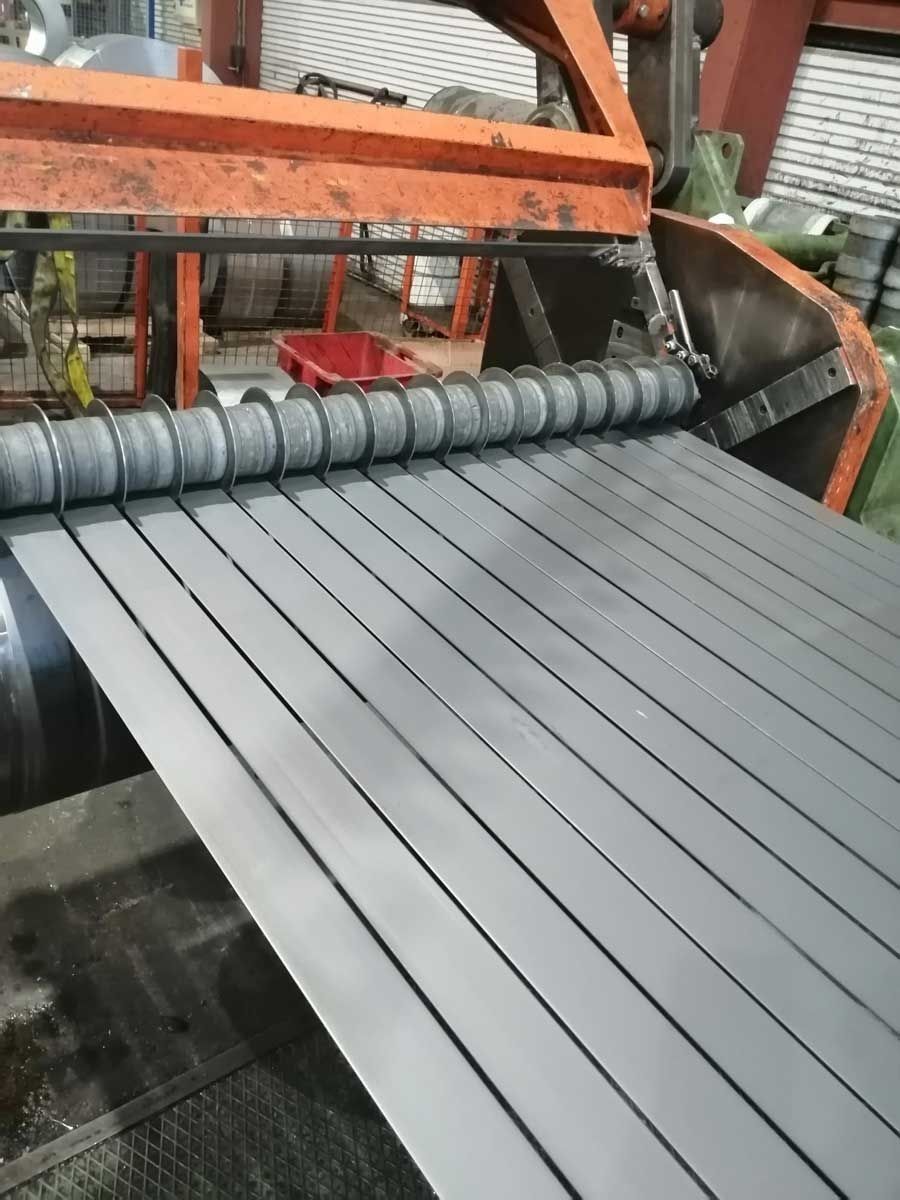 Cortadora de chapa de acero cortando tiras de una bobina grande en una fábrica. Maquinaria naranja y metálica.