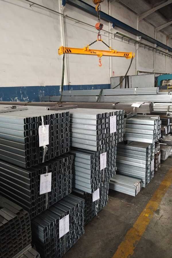 Pilas de vigas metálicas en forma de C en una fábrica. Una viga de elevación amarilla está suspendida encima.