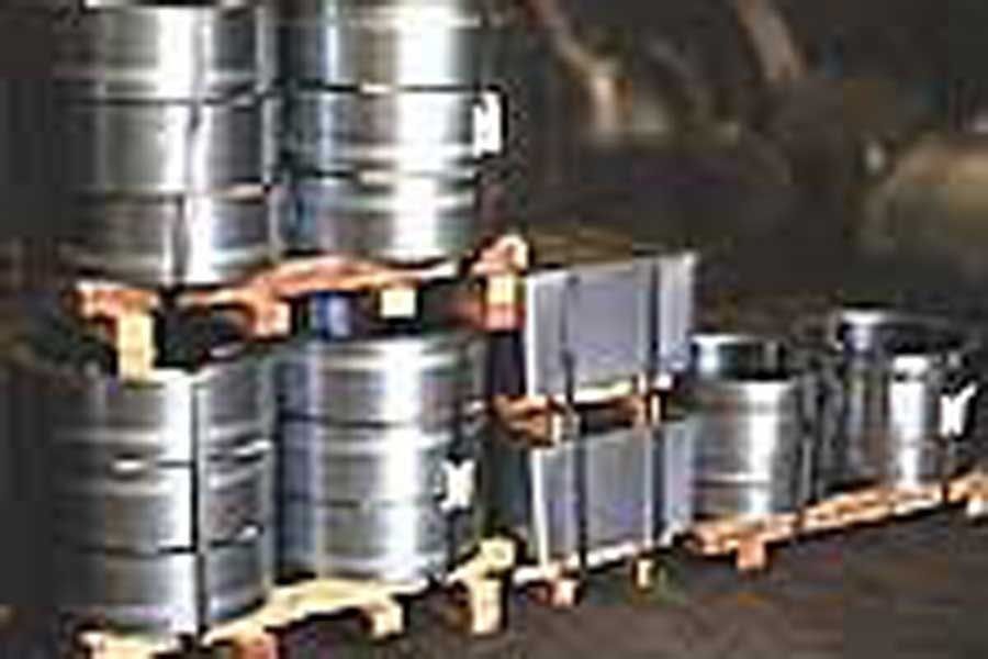 Pilas de barriles de metal brillante y piezas de metal rectangulares sobre paletas de madera en un almacén.