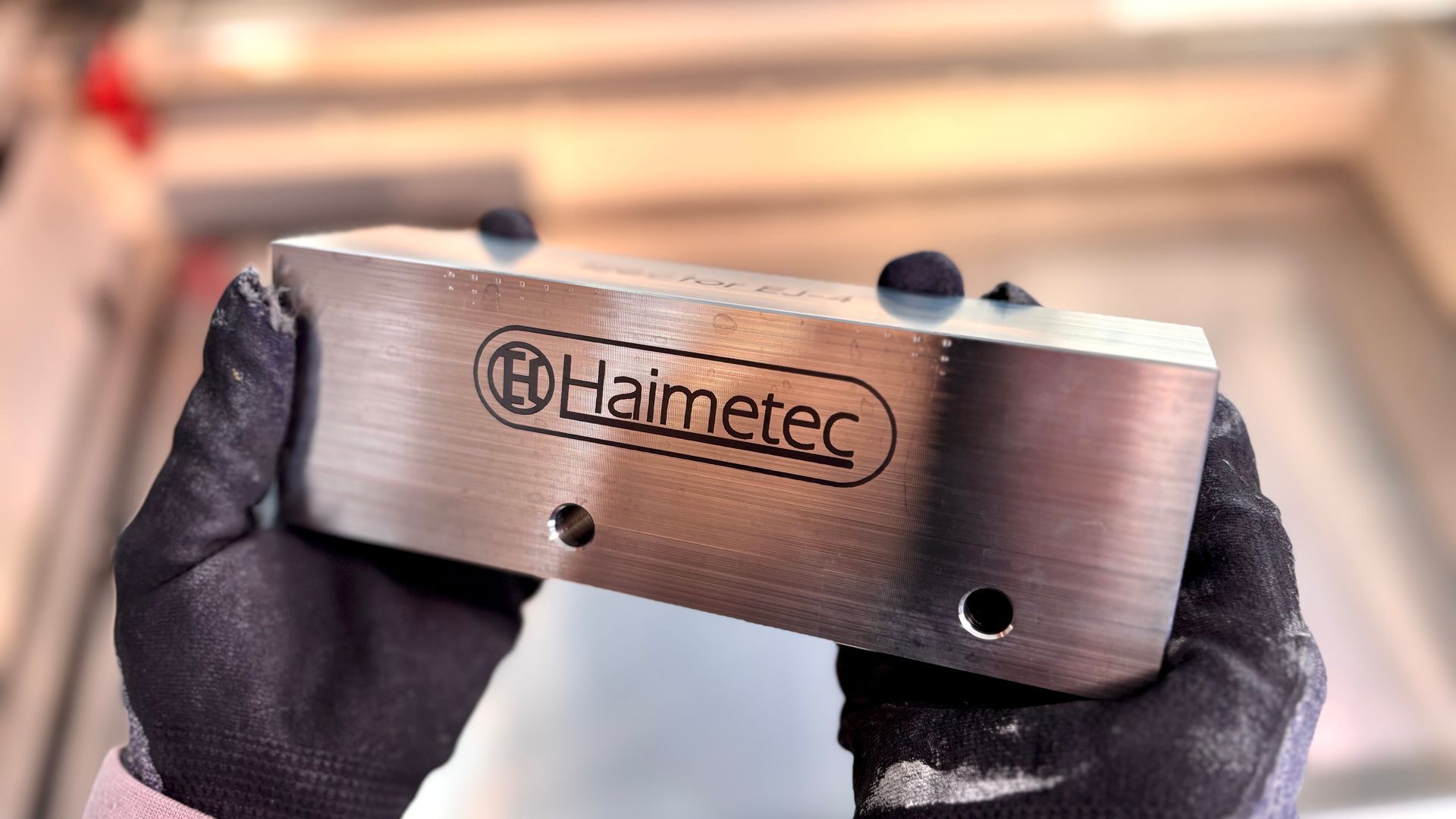 Käsineillä verhotut kädet pitelevät kahta reikää sisältävää Haimtec-merkkistä metallilevyä verstasympäristössä