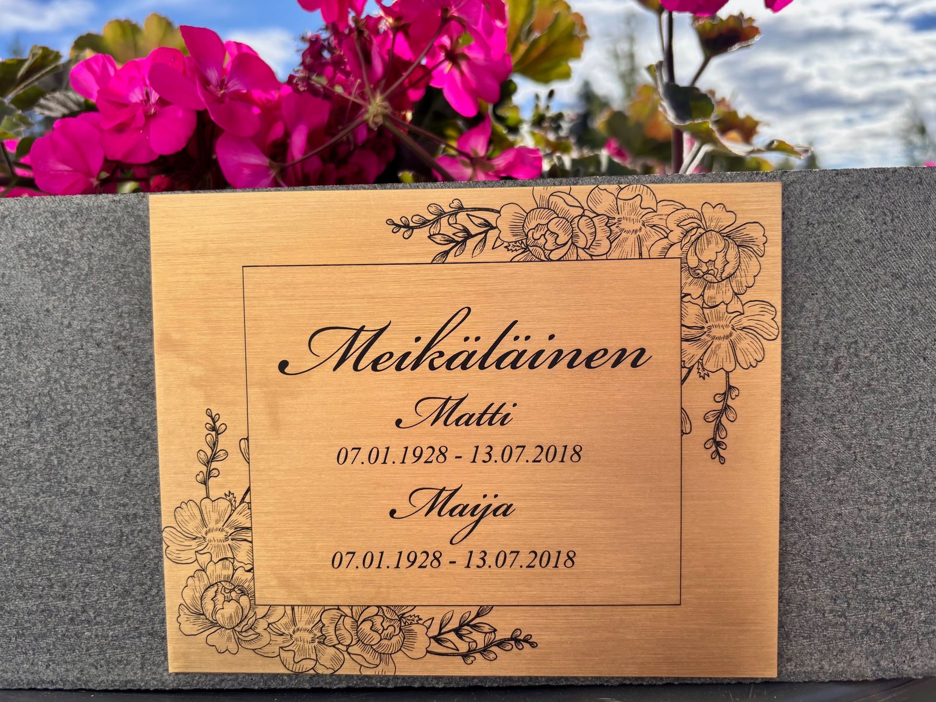 Kuparinen muistolaatta, johon on kaiverrettu nimiä, harmaata seinää vasten; yläpuolella vaaleanpunaisia kukkia