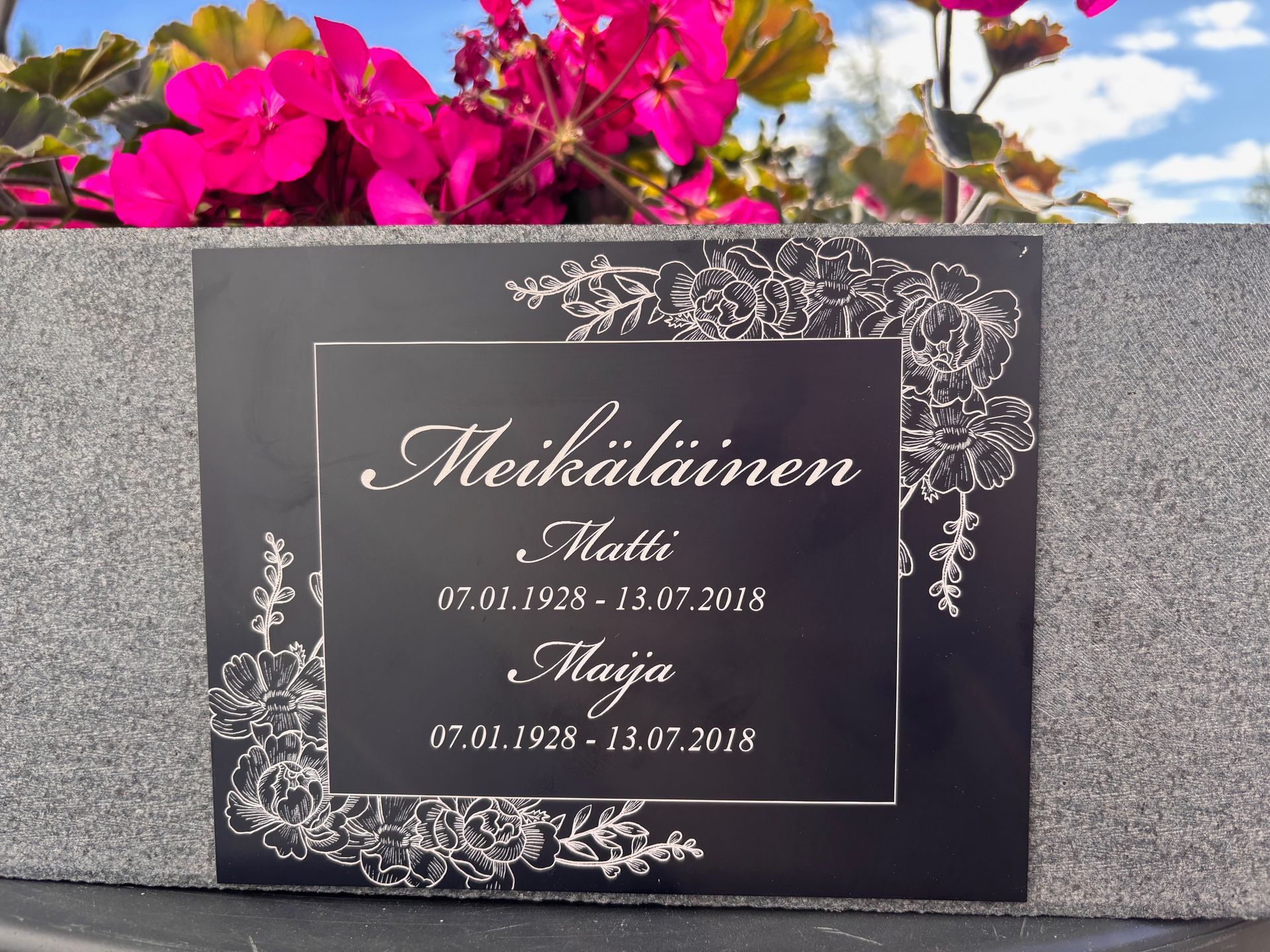 Muistolaatta, jossa on valkoista kaunokirjoitusta ja vaaleanpunaisia kukkia harmaata kivitaustaa vasten